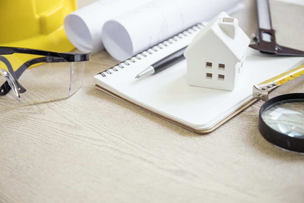 Small model house on a notebook with construction tools, safety glasses, and blueprints on a desk.